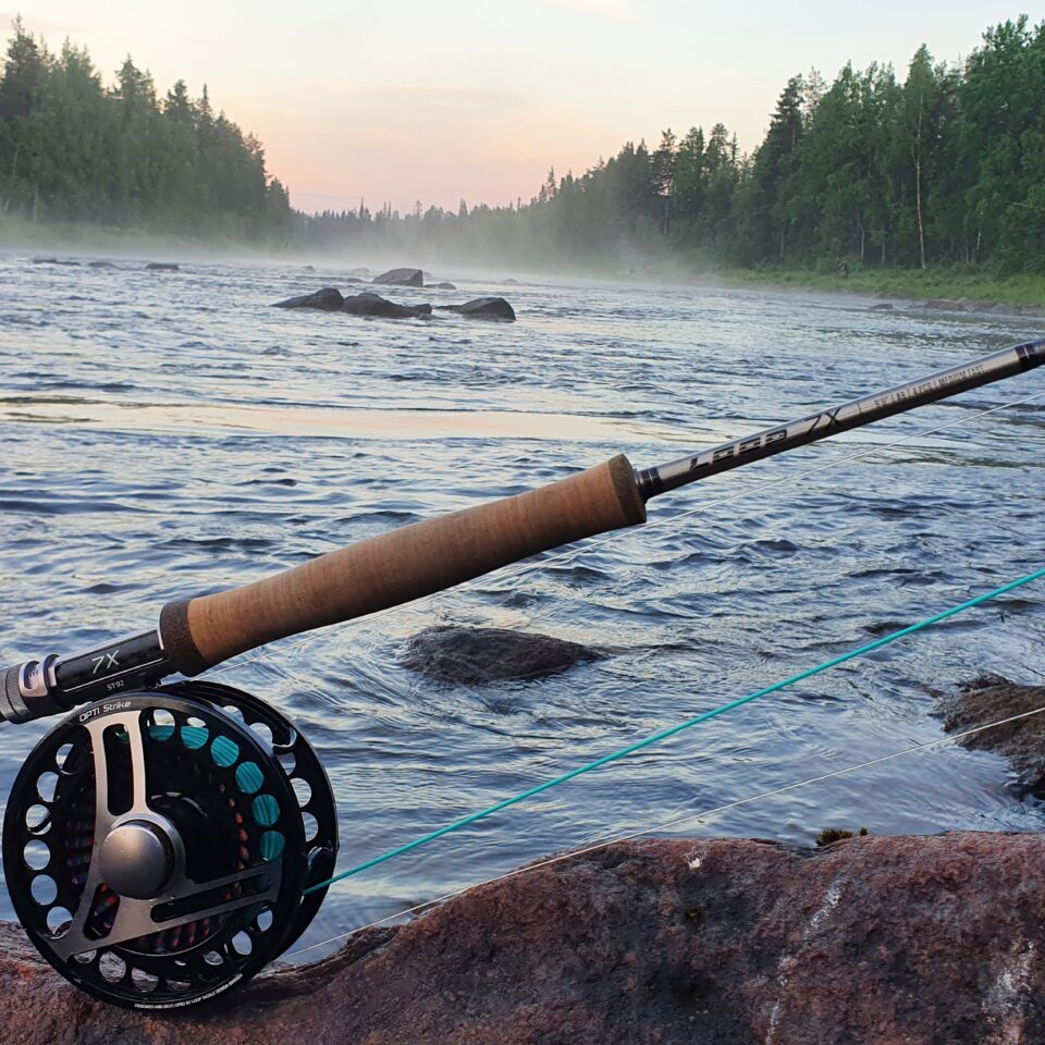 Ett fiskespö som ligger på en sten, i bakgrunden syns en älv med skog på sidorna.