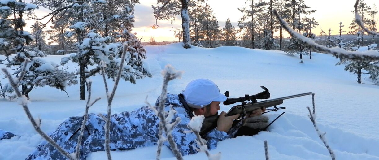 Person ligger i snö med kamouflerade kläder hållandes ett jaktgevär.