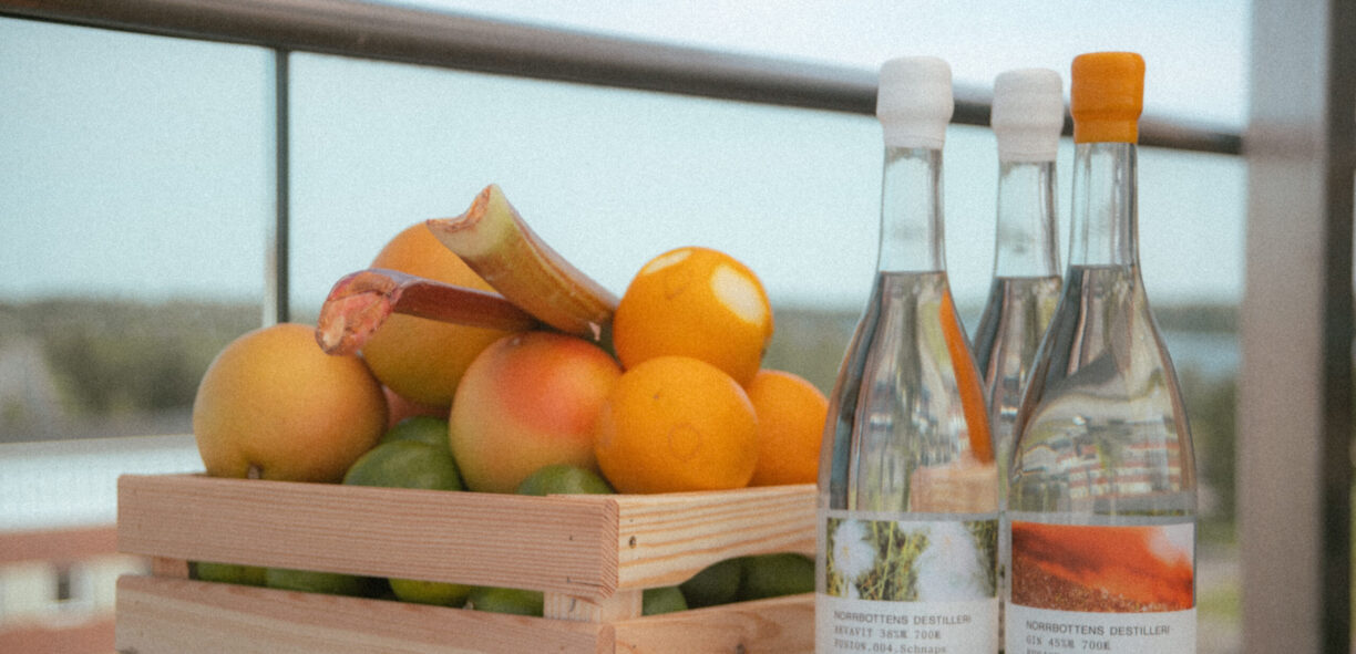 En trälåda med orangea frukter som står på en bardisk tillsammans med genomskinliga glasflaskor med dryck. Etiketterna på flaskorna är olika med text.