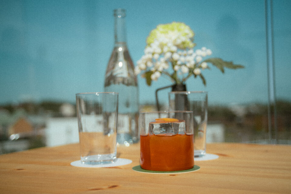 Ett träbord där det står glas med dryck samt en glasflaska. I bakgrunden skymtas skog och en blå himmel. En vit blomma i en vas står även på bordet.