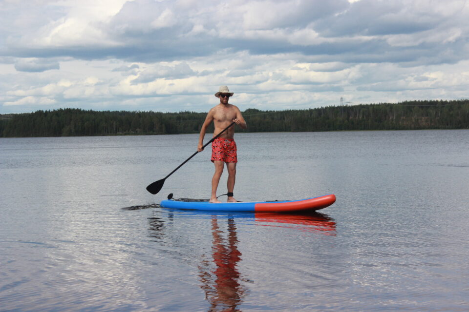 Person ståendes på en SUP, en stand-up-paddeling board, ute i vattnet med skog bakom och molntäckt himmel.