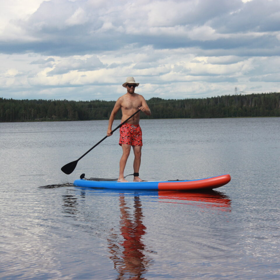 Person ståendes på en SUP, en stand-up-paddeling board, ute i vattnet med skog bakom och molntäckt himmel.