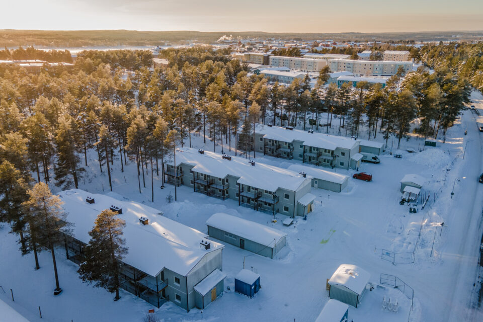 Flygbild av låga flerbostadshus som ligger naturskönt vid en tallskog men nära sjukhus med mera. Snö och sol.