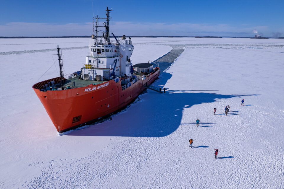 Stort rött fartyg som brutit en ränna i isen. Människor går på isen bredvid.