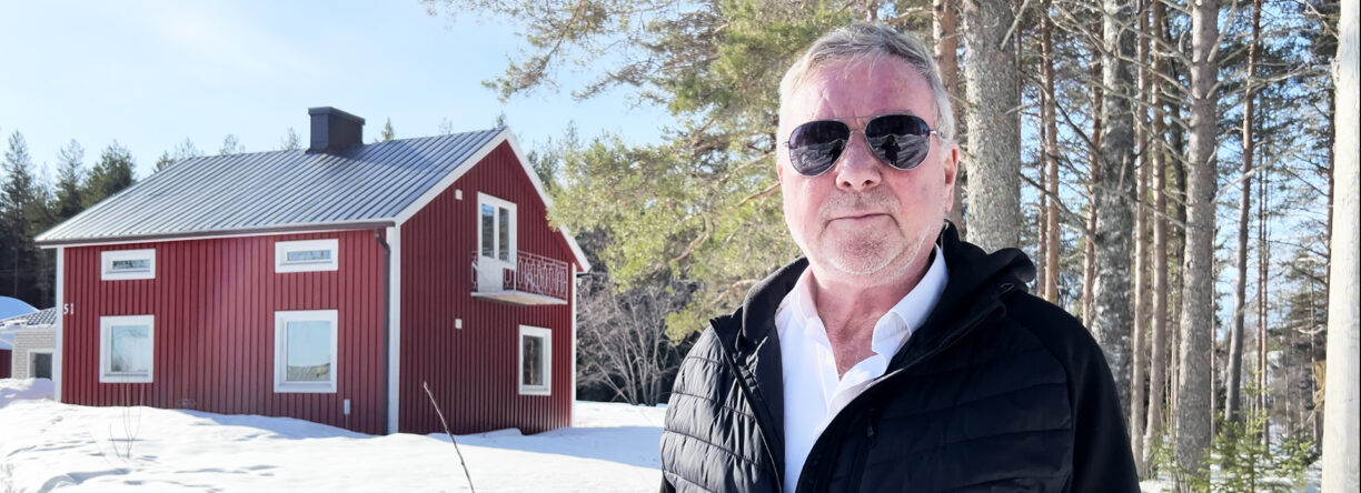 A man, Eddy Stam, is stnading in front of a red house. Snow on the ground and blue sky.
