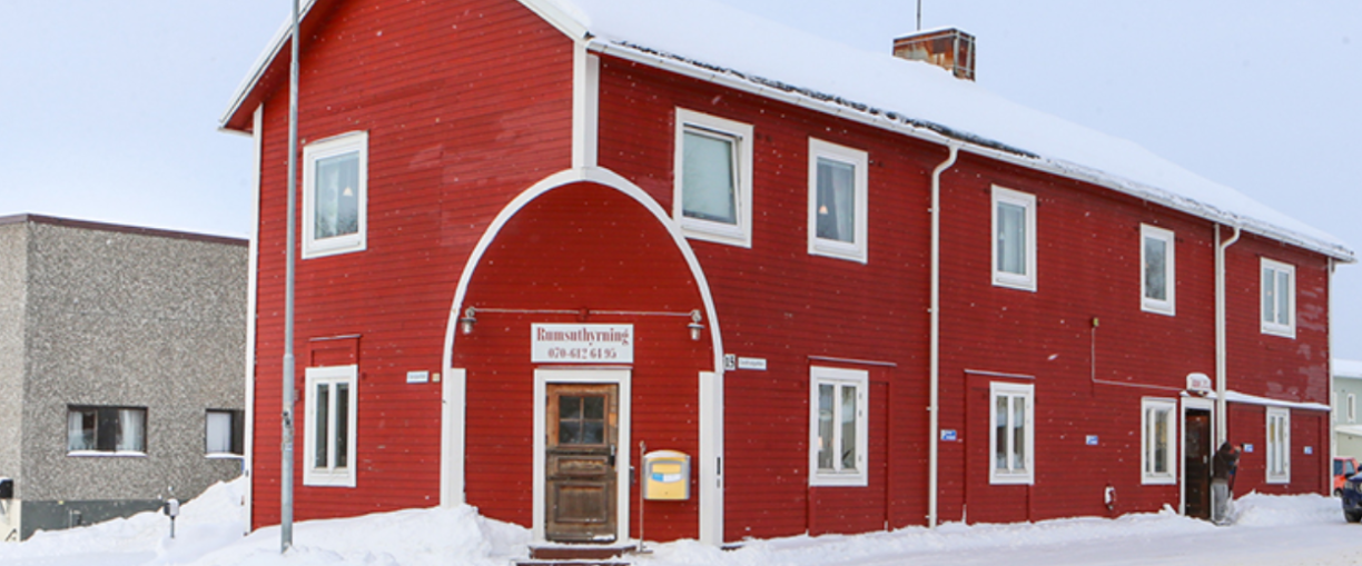Stort rött avlångt hus med två våningar. Huset har en brun dörr och snön ligger vit på marken framför huset.
