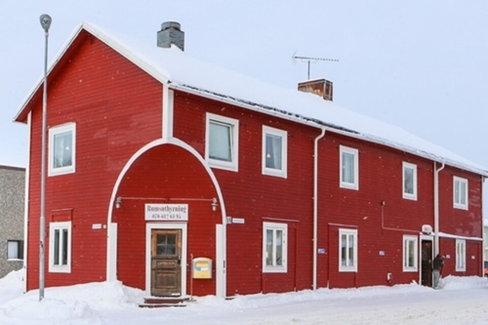 Stort rött avlångt hus med två våningar. Huset har en brun dörr och snön ligger vit på marken framför huset.