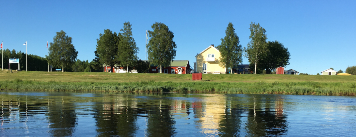 Sangisälven i förgrunden och ett gult hus samt tre mindre röda små hus står vid älvkanten. Tre flaggstänger syns till vänster i bild.