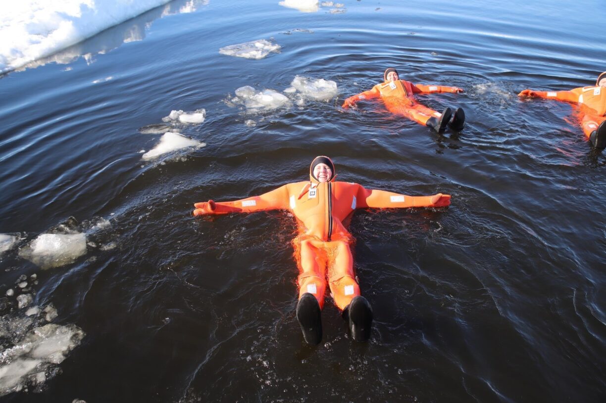 Tre personer badar isvak med överlevnadsdräkt, vattnet är mörkt och viss is syns bland människorna som badar.