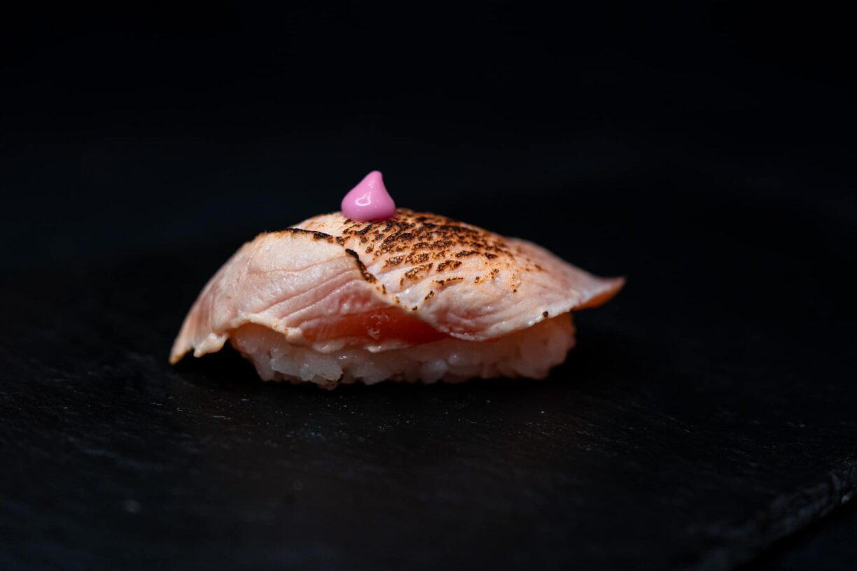 En sushibit med halstrad lax samt en klick lila sås. Bakgrunden är helt i svart, och sushibiten är fotad ganska nära.