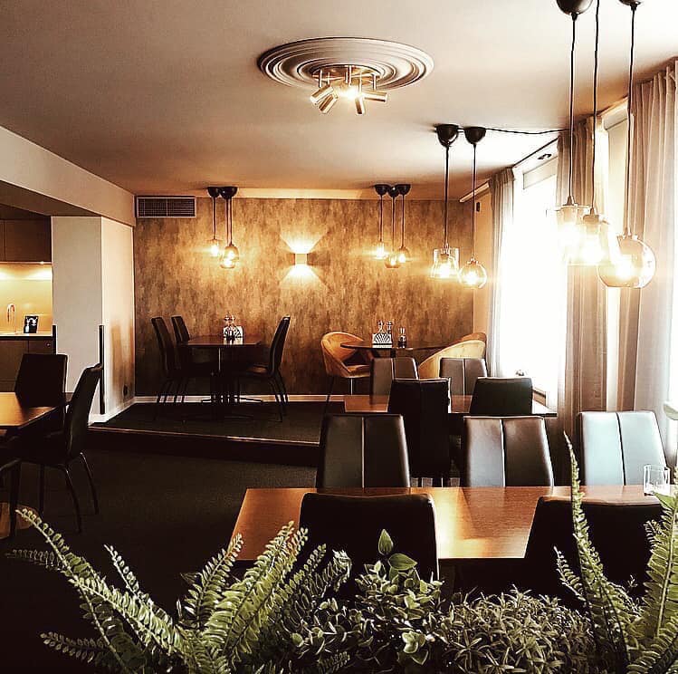 Bild tagen in ifrån restaurang Gamla Staden. På bilden syns gröns växter i förgrunden, bruna bord med svarta skinnstolar är placerade på ett mörkt golv. En brunmelerad tapetvägg syns längst bort i rummet, glaslampor hänger från taket.