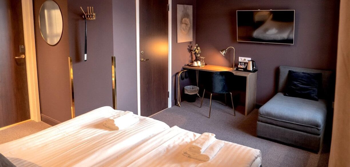 Ett hotellrum i dova lilabruna färger. en dubbelsäng med vita sängkläder i nedre kant och en skrivbord samt en utbäddbar fåtölj syns på bilden.