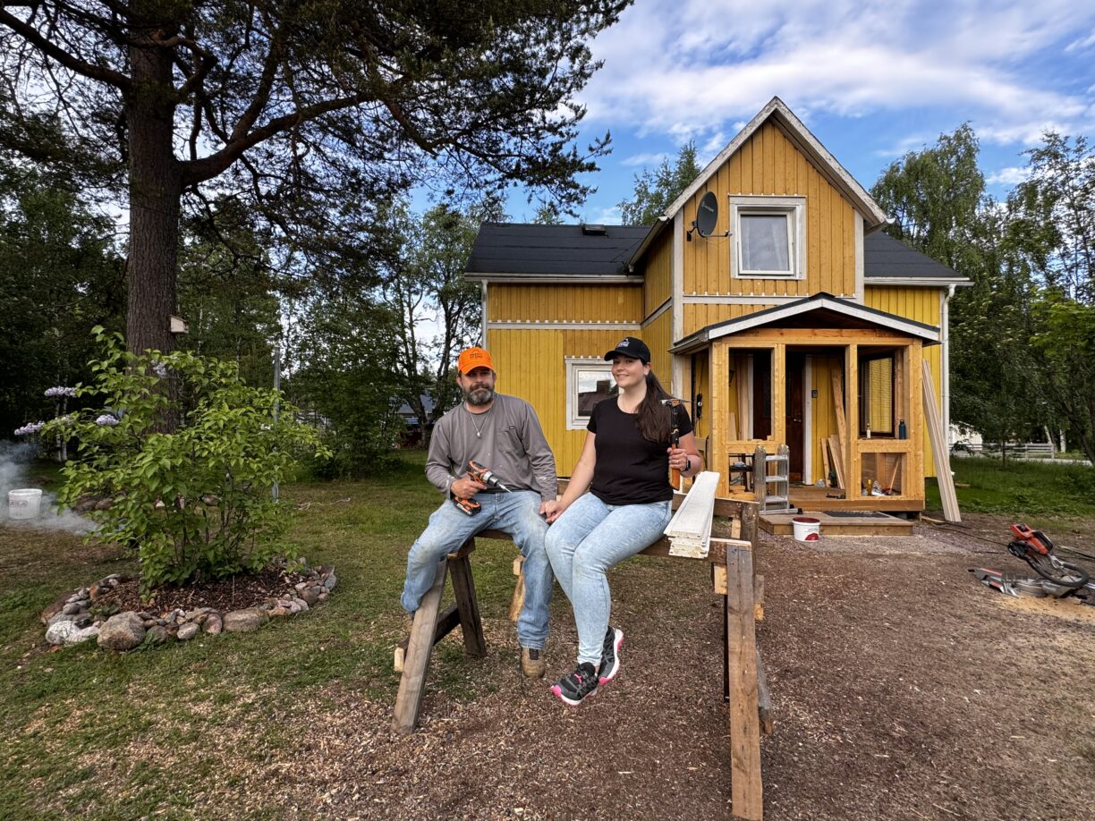 Mimi och Derrick sitter framför sitt gula hus. Till vänster syns lite grönska och ett träd.