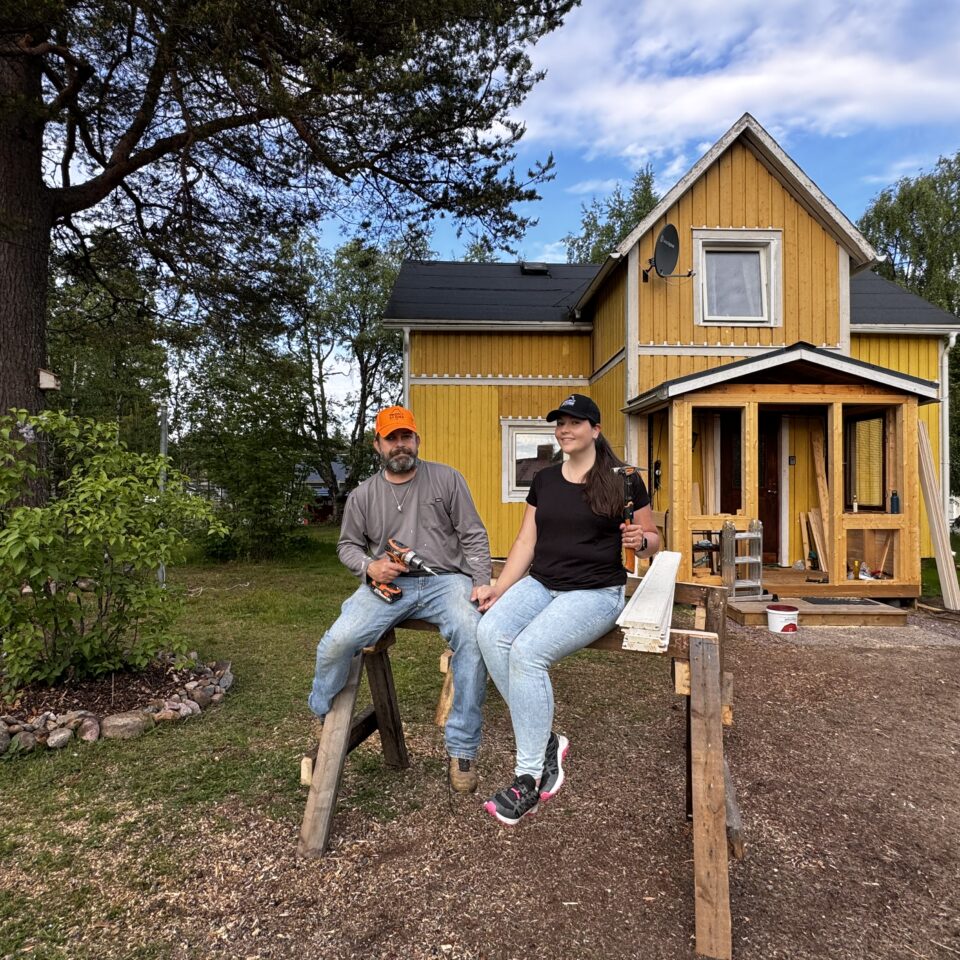 Mimi och Derrick sitter framför sitt gula hus. Till vänster syns lite grönska och ett träd.