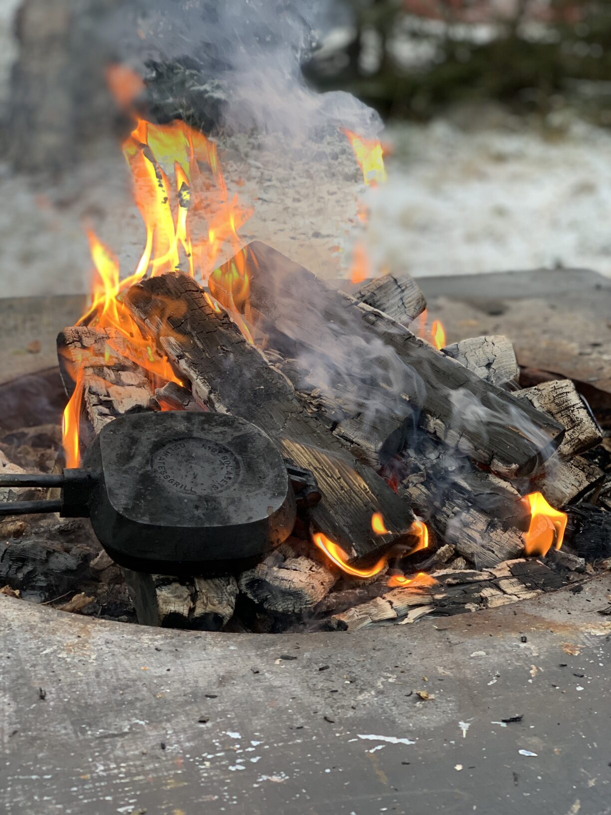 En brasa med gul-orange eldslågor i en grill-ring. Ett smörgåsjärn ligger på elden.