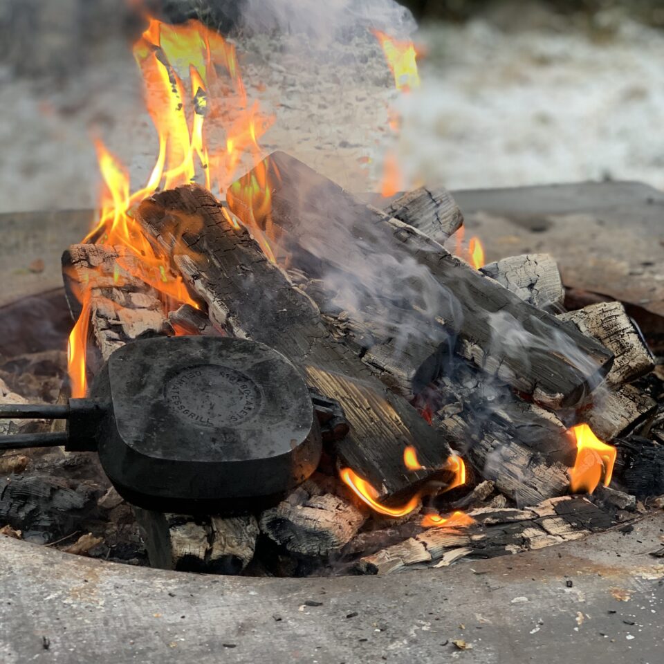 En brasa med gul-orange eldslågor i en grill-ring. Ett smörgåsjärn ligger på elden.