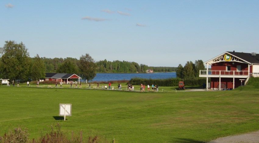 Grön gräsmatta, golfbana, med flera röda hus, människor som spelar golf. Mitt i bilden skymtar man blått vatten, Kalixälven. gröna lövträd i vänster kant och blå himmel i övre kant på bilden.