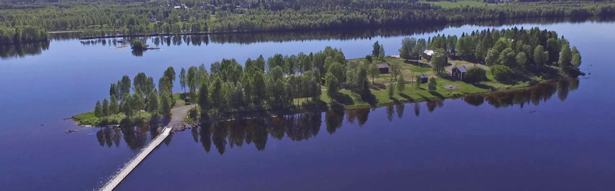 Vy över ön Vassholmen i Kalixälven. Grönskande ytor med träd och röda byggnader. Vatten runt om.