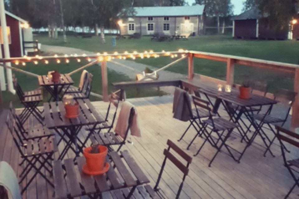 En träaltan med cafébord och stolar i förgrunden, i bakgrunden syns grönt gräs och en grånad lada och gröna träd.