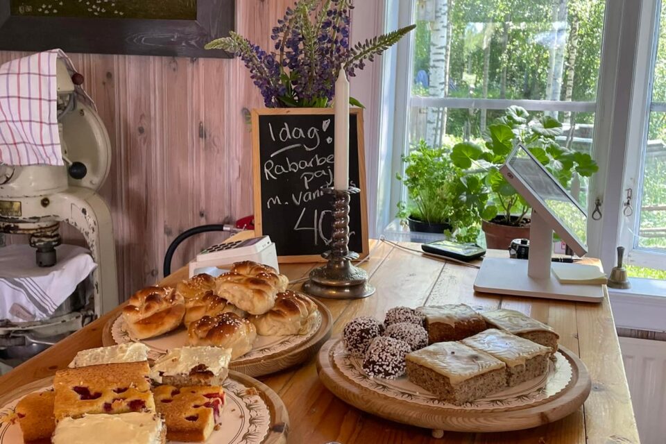 Ett uppdukat bord med hembakt fika. Ett fönster med en vit gardin och blommor syns baka bordet.