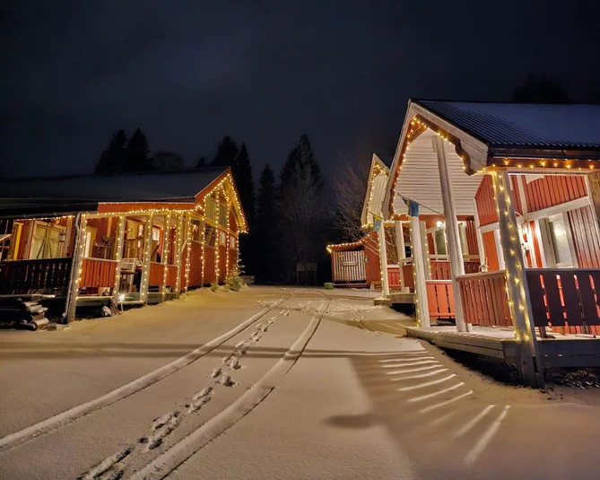Röda stugor med vita knutar på rad med julbelysning. Marken är täckt med snö.