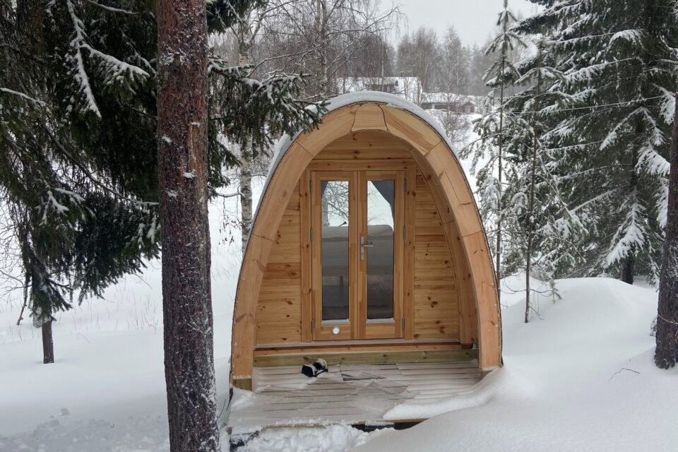 En liten stuga i trä med två glasdörrar i mitten, stugan har ett rundat tak som går från ena sidan till den andra. Stugan står omgiven av snö och granar.