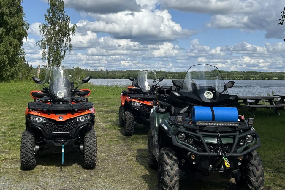 Tre fyrhjulingar som står på en gräsmatta med lite grus på.I bakgrunden syns älven och ovanför den en blå himmel med vita moln. Vid älvkanten syns några träd.