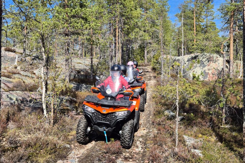 Två röda fyrhjulingar som kör på en skogsväg. Tallar, granar runt om, blå himmel ovanför.