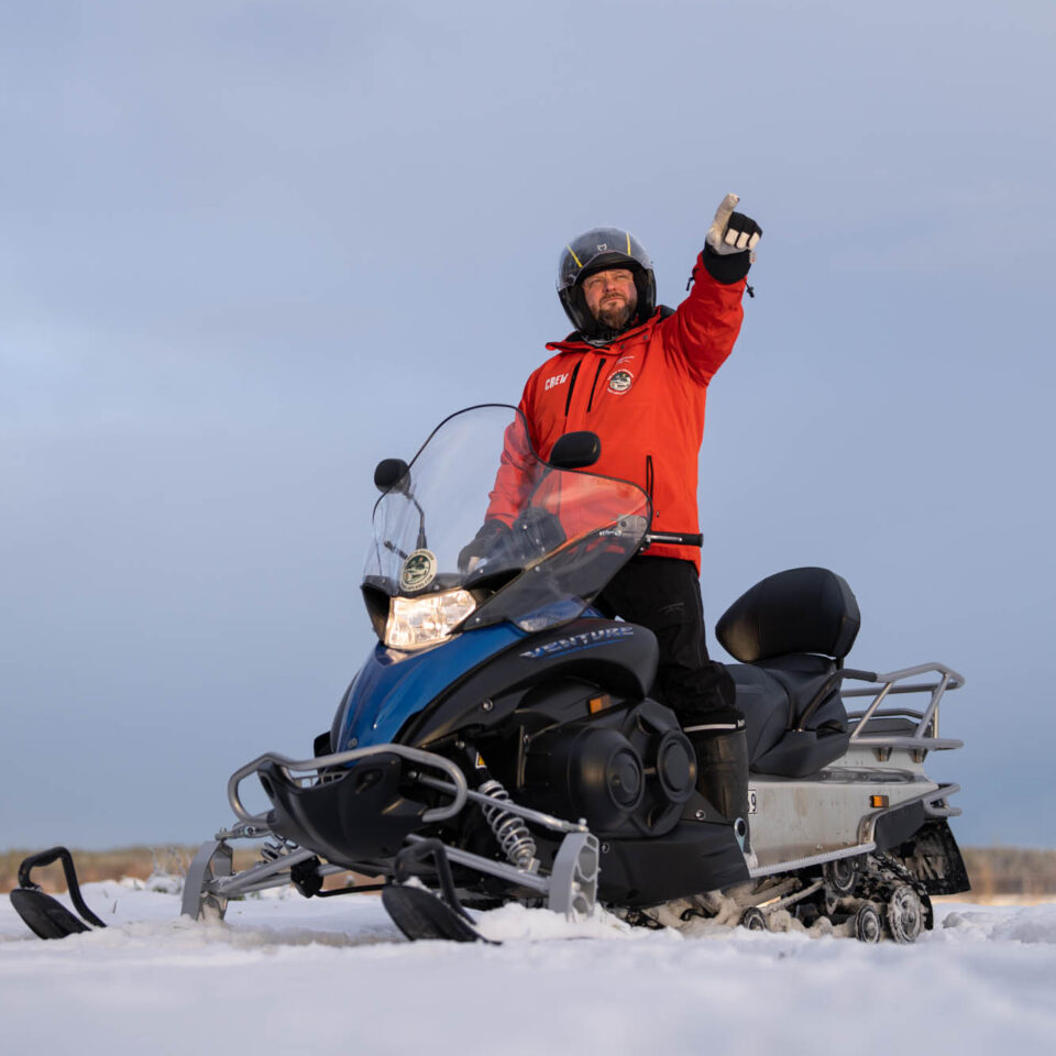 Vit snötäckt mark med en svart och blå skoter och en man med svarta byxor och orange jacka syns på skotern. Han pekar mot himlen som är gråblå.