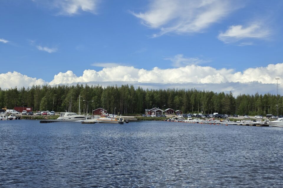 Vy-bild över båthamn från vattnet. Granskog och blå himmel med vita moln i bakgrunden.